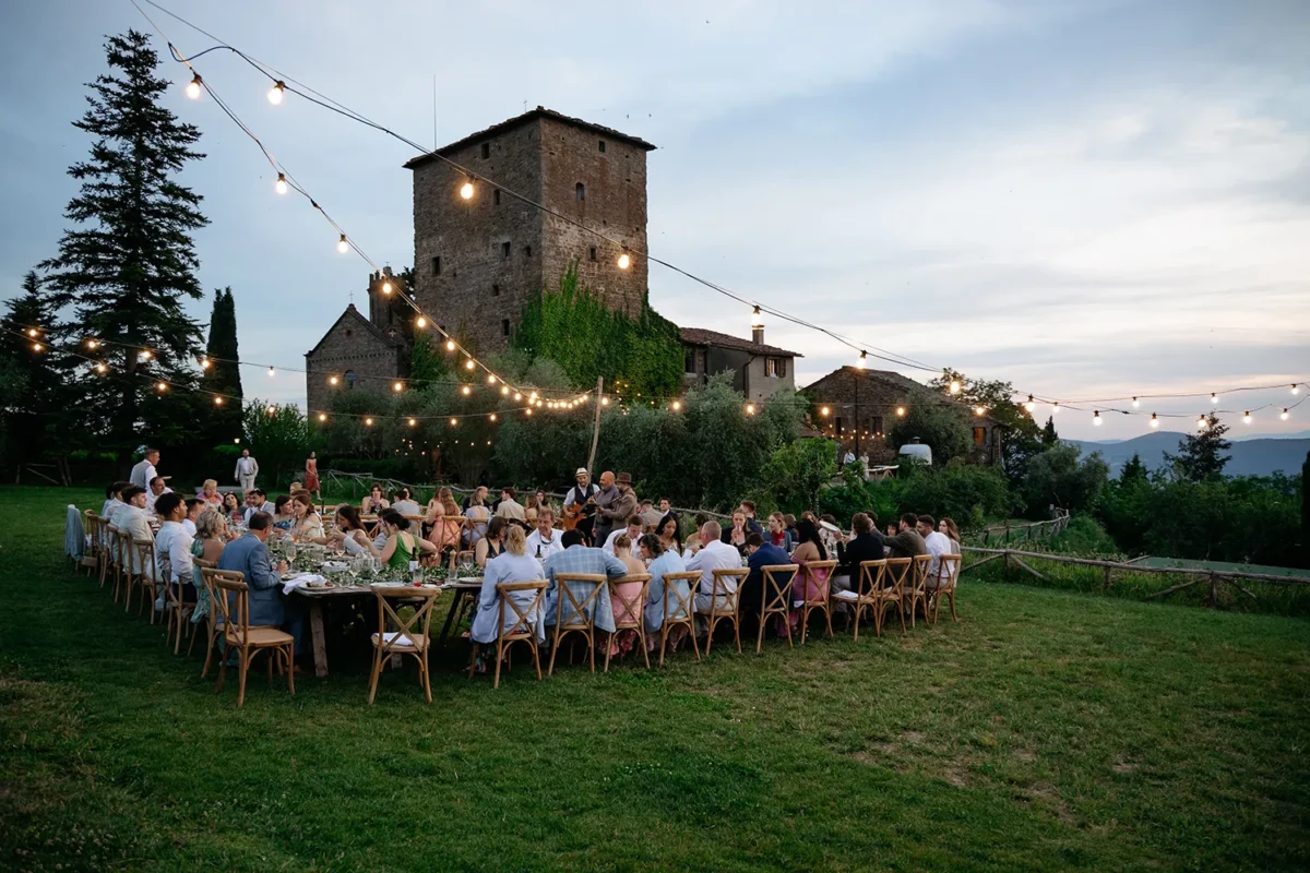 Long wedding dinner table on the lawn under string lights, with a stone tower behind, Tuscany